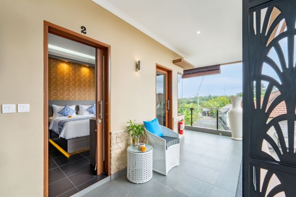 Spacious tropical villa balcony with wicker lounge chair and table, open door to cozy bedroom, overlooking lush Balinese greenery.